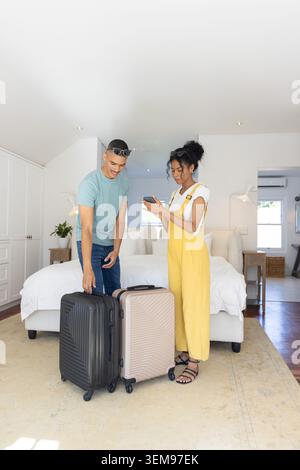 Couple diversifié debout dans la chambre avec des bagages, femme en salopette jaune vérifiant le téléphone Banque D'Images