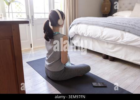 Femme assise sur les jambes croisées sur le tapis de yoga dans la chambre face aux doubles portes en verre avec smartphone sur le tapis Banque D'Images
