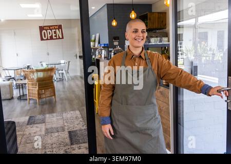 Femme ouvrière de café portant un tablier vert gris tenant la porte vitrée, souriant au comptoir local, espace de copie Banque D'Images
