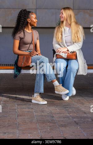 Diverses amies féminines assis sur un banc en bois à plaza, souriant et tenant des sacs à main bruns Banque D'Images
