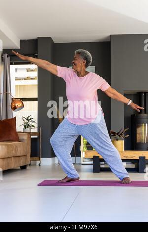Femme afro-américaine senior pratiquant le yoga debout, équilibre sur tapis violet dans le salon Banque D'Images