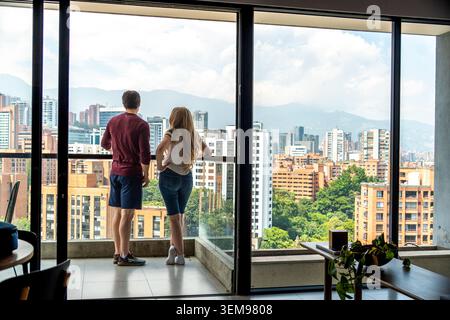 Deux personnes se tiennent sur un balcon d'appartement moderne, donnant sur un paysage urbain animé de grands bâtiments, des balustrades en verre, et des montagnes lointaines baignées dans d Banque D'Images