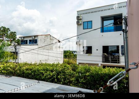 Les maisons blanches de style appartement avec balcons, fenêtres, climatiseurs et fils électriques visibles se tiennent derrière une dense haie verte sous un fourneau Banque D'Images