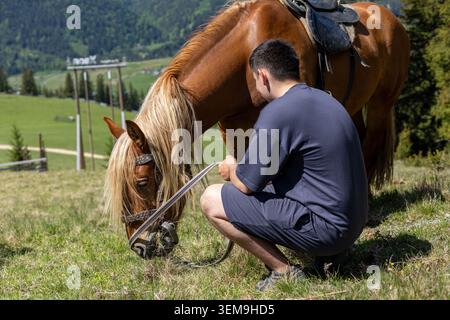 Homme se reposant à côté d'un cheval sellé dans les montagnes. Homme accroupi à côté d'un cheval selle brune pâturant sur une colline alpine avec vallée verdoyante et Banque D'Images