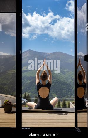 Femme faisant du yoga avec vue sur la montagne. Femme pratiquant le yoga sur un balcon à une porte ouverte avec des montagnes vertes panoramiques, un ciel bleu et des nuages au-delà Banque D'Images
