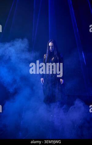 Femme adulte avec de longs cheveux noirs portant une robe gothique sombre se tient au milieu d'un épais brouillard bleu sous un éclairage dramatique, créant une atmosphère mystérieuse et étrange Banque D'Images