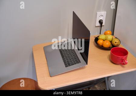 Ordinateur portable argenté élégant ouvert sur le bureau d'angle en bois clair à côté d'un bol de fruits noirs avec des poires et des oranges et une tasse de café rouge, mis contre le mur gris Uni wi Banque D'Images