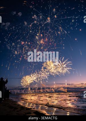 Spectaculaire feu d'artifice doré illuminant le ciel nocturne et se reflétant sur les vagues de la mer lors d'un événement estival côtier en Europe. Banque D'Images