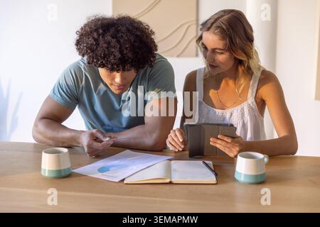 Couple diversifié assis à la table à manger en bois examinant les reçus et tablette avec des tasses sarcelle Banque D'Images