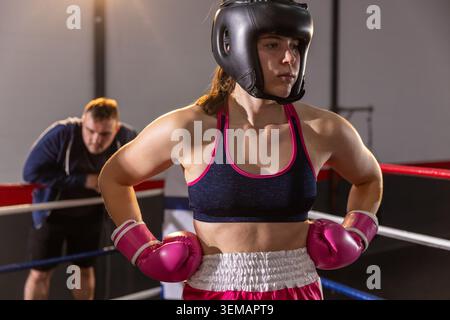 Boxeur féminin avec entraîneur posant les mains sur les hanches dans l'anneau portant des gants roses et un couvre-chef Banque D'Images