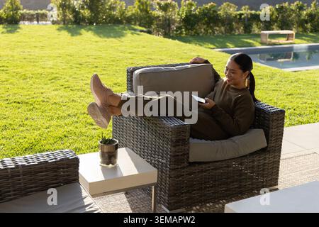 Femme asiatique assise dans une chaise en osier sur le patio à l'aide d'un smartphone dans les joggers d'olive, espace de copie Banque D'Images