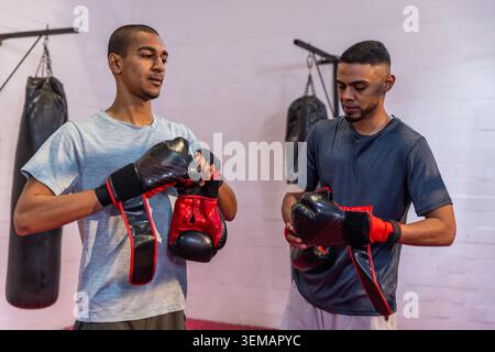Deux hommes ajustant des gants rouges-noirs et des enveloppements dans des vêtements de sport près de sacs suspendus dans la salle de boxe Banque D'Images