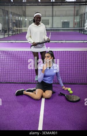 Diverses amies féminines assises sur un terrain violet tenant des pagaies et des boules vertes néon près du filet Banque D'Images