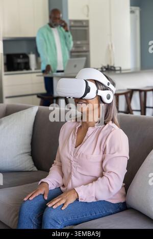 Couple senior à la maison, femme portant un casque VR blanc sur un canapé gris, homme utilisant un smartphone Banque D'Images