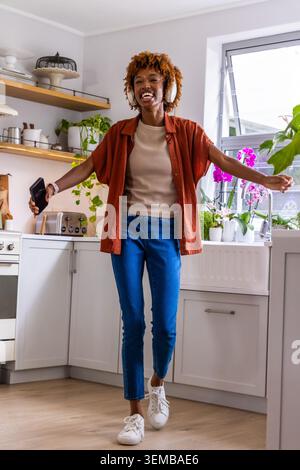 Femme afro-américaine dansant dans la cuisine, écoutant sur un casque sans fil et tenant un smartphone Banque D'Images