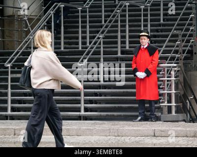 Une femme d'affaires élégamment habillée passe devant l'emblématique bâtiment Lloyd's dans la City de Londres, au Royaume-Uni, tandis qu'un serveur traditionnel Lloyd's, servant de sécurité, se tient près de l'entrée vêtu d'un haut-de-forme et d'un manteau cramoisi Banque D'Images