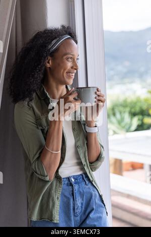 Femme afro-américaine tenant une tasse tout en regardant à travers la porte coulissante en verre au balcon, espace de copie Banque D'Images