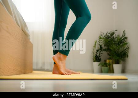 Femme debout sur le tapis de yoga jaune se préparant pour l'entraînement à la maison dans la chambre. Équilibre féminin actif et étirement pour un mode de vie sain. Dame restant en forme grâce à la pratique régulière de fitness à domicile Banque D'Images
