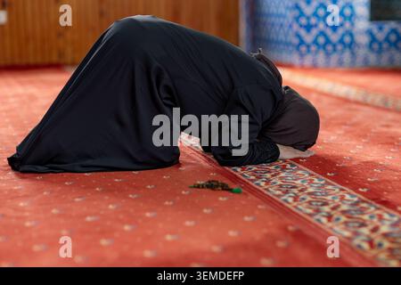 Femme musulmane en hijab noir effectue la prière sujood à l'intérieur d'une mosquée, s'inclinant sur un tapis rouge à motifs dans un espace sacré calme, exprimant la foi et le développement Banque D'Images
