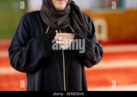 Gros plan d'une femme musulmane en abaya et hijab debout dans une mosquée avec les mains pliées en prière, montrant la foi, la dévotion et la réflexion spirituelle tranquille Banque D'Images