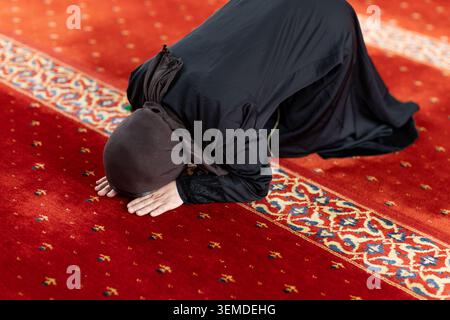 Femme musulmane en hijab noir effectue la prière sujood à l'intérieur d'une mosquée, s'inclinant sur un tapis rouge à motifs dans un espace sacré calme, exprimant la foi et le développement Banque D'Images