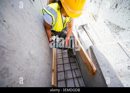 Ouvrière de construction en casque de sécurité et gilet de sécurité perce dans le béton à l'intérieur d'une tranchée étroite avec barre d'armature, se concentrant sur les travaux de rénovation et le chantier Banque D'Images