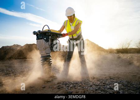 Travailleuse de la construction portant un casque de sécurité et un gilet de sécurité utilise un compacteur à plaque vibrante sur du gravier sur un chantier poussiéreux, rétroéclairé par un soleil éclatant et s Banque D'Images