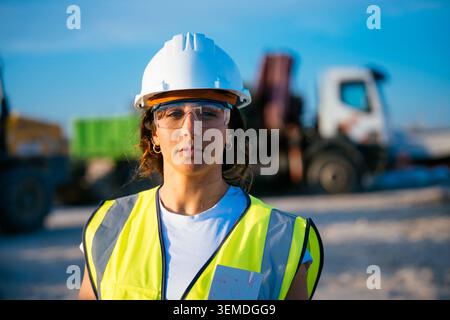 Travailleuse de construction confiante portant un casque de sécurité, des lunettes de sécurité et un gilet réfléchissant sur un chantier actif, avec des machines floues dans le backgrou Banque D'Images