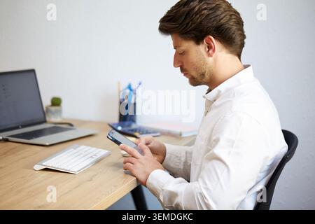 Un homme professionnel en chemise blanche travaille sur un bureau en bois, à l'aide d'un smartphone. Un ordinateur portable et des fournitures de bureau l'entourent, capturant le travail moderne e Banque D'Images