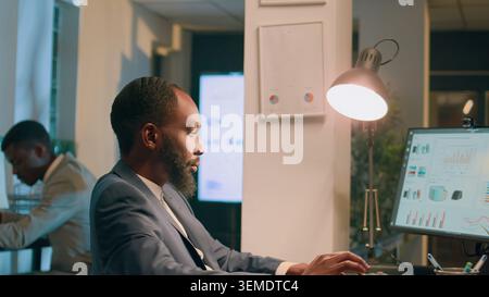 Hommes d'affaires afro-américains assis à leur bureau, regardant sur des graphiques financiers, imputant des données sur ordinateur pendant le quart de nuit. Collègues résolvant des tâches de projet d'entreprise du jour au lendemain avant la date limite Banque D'Images
