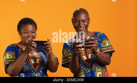 Couple afro-américain jouant à des jeux vidéo mobiles sur caméra, s'amusant avec tournoi en ligne sur fond orange. Joueurs heureux appréciant le concours Internet ensemble. Banque D'Images