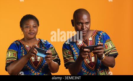 Couple afro-américain jouant à des jeux vidéo mobiles sur caméra, s'amusant avec tournoi en ligne sur fond orange. Joueurs heureux appréciant le concours Internet ensemble. Banque D'Images
