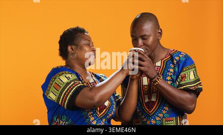 Couple ethnique appréciant l'arôme de boisson de café frais, sentant la boisson de caféine tandis qu'ils posent sur fond orange. Jeunes partenaires dans une tenue traditionnelle aimant le parfum de rafraîchissement. Banque D'Images