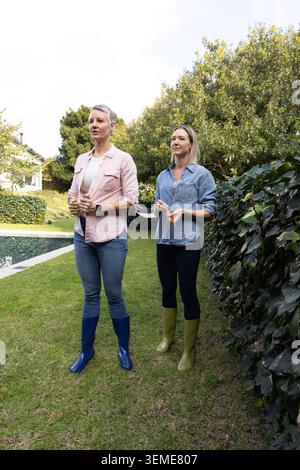 Amies d'âge moyen debout sur la cour près de la piscine portant des bottes hauteur genou tenant un outil orange Banque D'Images