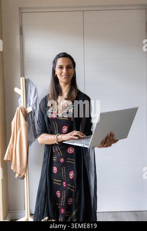 Femme indienne debout tenant un ordinateur portable argenté à la maison portant une robe imprimée noire par porte-manteau Banque D'Images