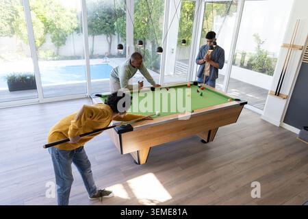 Divers amis masculins jouant sur la table en feutre vert tenant des repères dans la salle de jeux moderne près des portes vitrées Banque D'Images