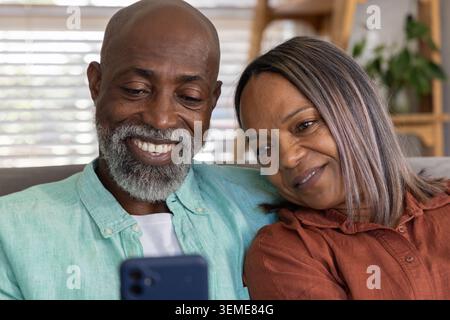 Couple mature diversifié assis sur le canapé à la maison, penchés ensemble tout en regardant smartphone Banque D'Images