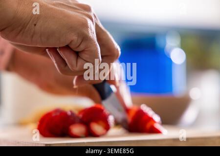 Main femelle mature saisissant couteau d'office tranchant des fraises sur planche de bois dans la cuisine, tasse floue Banque D'Images