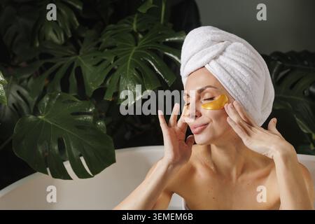 Une femme se détend dans une salle de bain sereine, en appliquant des patchs oculaires dorés tout en profitant d'un moment d'auto-soin entouré de plantes vertes luxuriantes. Banque D'Images