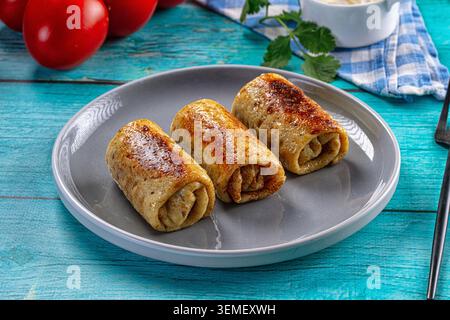 Trois crêpes frites farcies de viande sur assiette grise. Table en bois bleu. Tomates rouges. Persil. Bol blanc. Serviette à carreaux bleue. Fourche noire. Rustique Banque D'Images