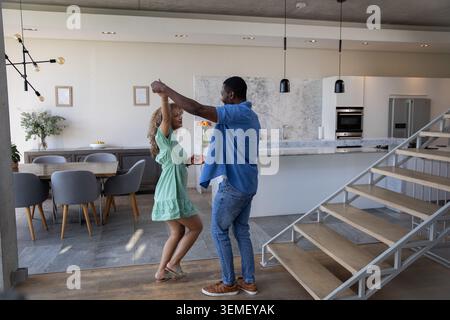 Couple diversifié dansant ensemble dans la cuisine ouverte près de l'îlot de marbre et escalier flottant Banque D'Images