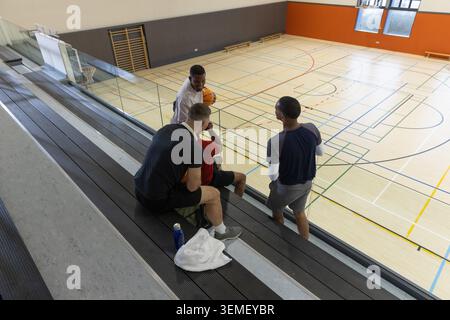 Divers coéquipiers masculins dans l'équipement de sport parlant tout en étant assis sur des gradins à la salle de gym de l'école avec balle Banque D'Images