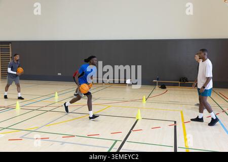 Divers coéquipiers masculins pratiquant l'exercice dribble sur un terrain de gymnastique avec des ballons de basket orange et des cônes Banque D'Images