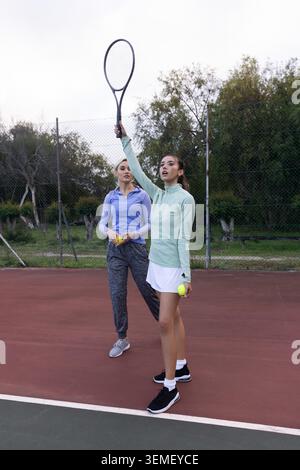 Deux femmes sur un court de tennis rouge, joueur de devant en raquette de levage de top menthe, partenaire tenant des balles Banque D'Images