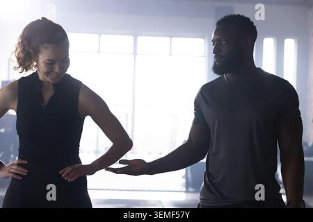 Couple diversifié debout dans le gymnase par de grandes fenêtres, en vêtements de sport, offrant la main près de l'équipement Banque D'Images