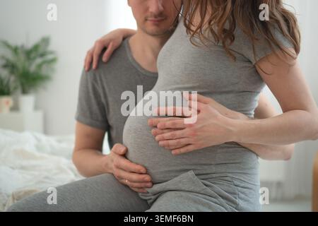 Femme enceinte et mari assis sur le lit sentant bébé bouger ensemble dans un moment intime. S'attendre à ce que la mère embrasse partenaire tout en expérimentant bébé coup de pied dans une chambre confortable. Maman-to-be partageant romantique Banque D'Images