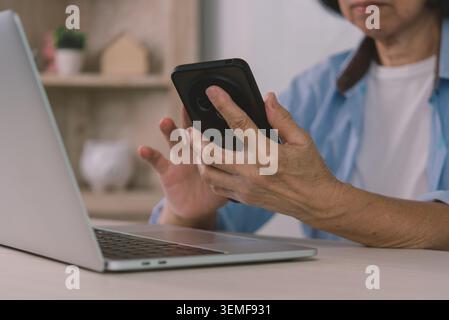 Gros plan de mains de femme senior tenant smartphone pour l'authentification à deux facteurs 2FA tout en travaillant sur ordinateur portable. Cybersécurité des personnes âgées, banque en ligne ve Banque D'Images