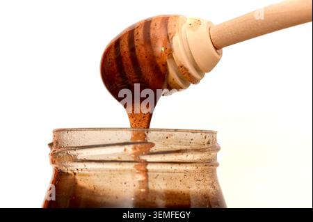 Gros plan de la tartinade au chocolat et aux noisettes coulant d'une trempette de miel en bois dans un bocal en verre sur fond blanc. Banque D'Images