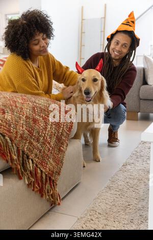 Couple sur le canapé à la maison, caressant Golden retriever portant un bandeau de corne rouge, homme portant un chapeau de citrouille Banque D'Images