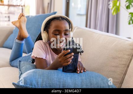 Jeune femme afro-américaine allongée sur un canapé, portant un casque crème et tenant un smartphone souriant Banque D'Images